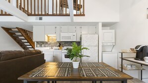 Una cocina moderna con armarios blancos, un microondas y un refrigerador. Una escalera de madera conduce a un nivel superior.