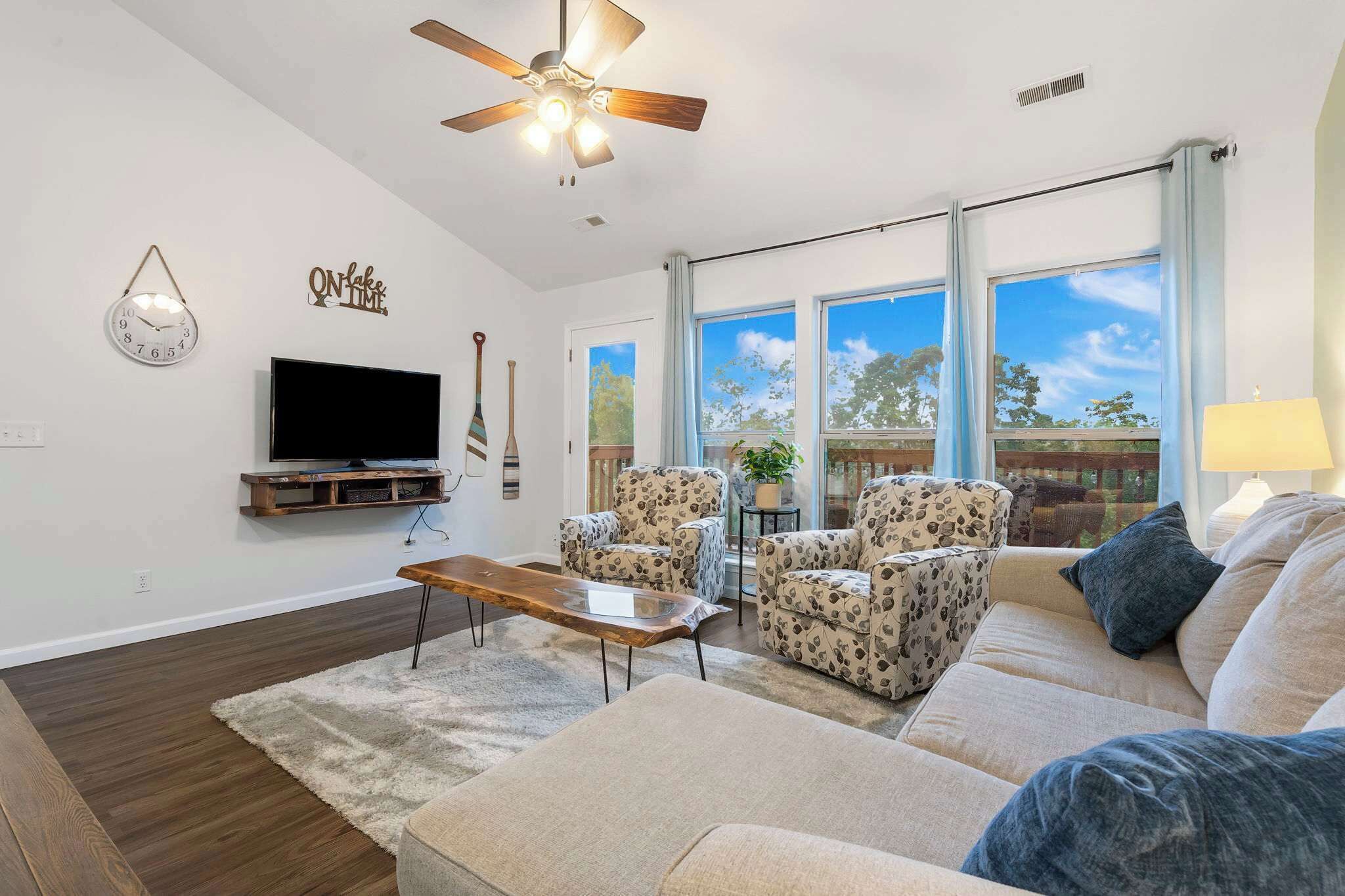 A living room with a flat-screen TV, a ceiling fan, a coffee table, two patterned armchairs, a sofa, and a window with a view of trees.
