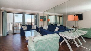 Ein modernes Wohnzimmer mit einem blauen Sofa, einem gemusterten Sessel und einem Glastisch. Ein großes Spiegelbild des Raumes und ein Blick auf den Ozean durch die Schiebetüren.