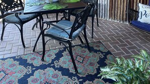 Eine überdachte Terrasse mit einem für vier Personen gedeckten Tisch, umgeben von Metallstühlen. Eine gewebte Leuchte hängt über dem Tisch, und eine Texas-Flagge ist an der Wand zu sehen.