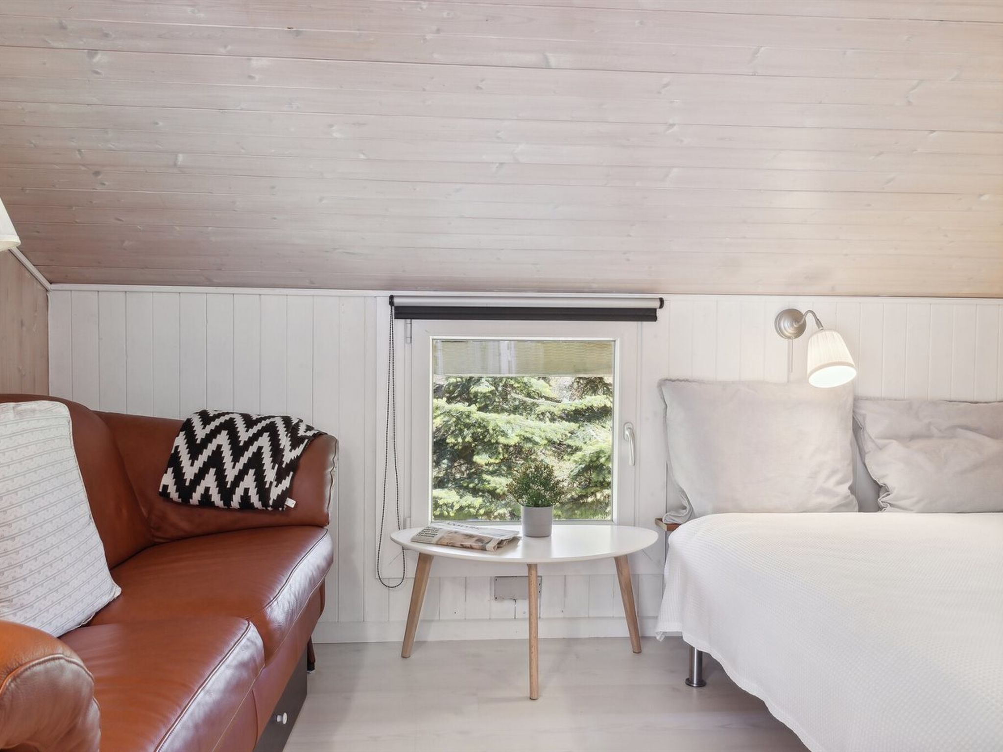 Une pièce chaleureuse avec un canapé en cuir marron, un lit blanc, une petite table ronde sur laquelle repose un magazine, et une fenêtre donnant sur un paysage verdoyant.