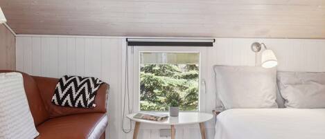 Une pièce chaleureuse avec un canapé en cuir marron, un lit blanc, une petite table ronde sur laquelle repose un magazine, et une fenêtre donnant sur un paysage verdoyant.