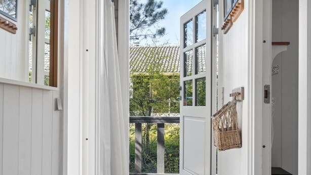 Un couloir étroit aux murs blancs, une porte en bois et une vue sur un balcon doté d’un toit en tuiles et bordé de verdure.