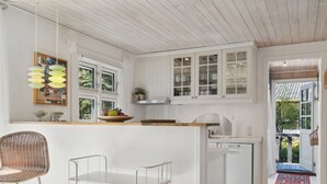 Une cuisine moderne avec des meubles blancs, un plan de travail en bois et un lave-vaisselle encastré. On y trouve une chaise en osier et un tabouret de bar en métal.