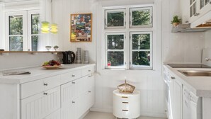 Une cuisine moderne avec des meubles blancs, une grande fenêtre et un panier de fruits posé sur le plan de travail.