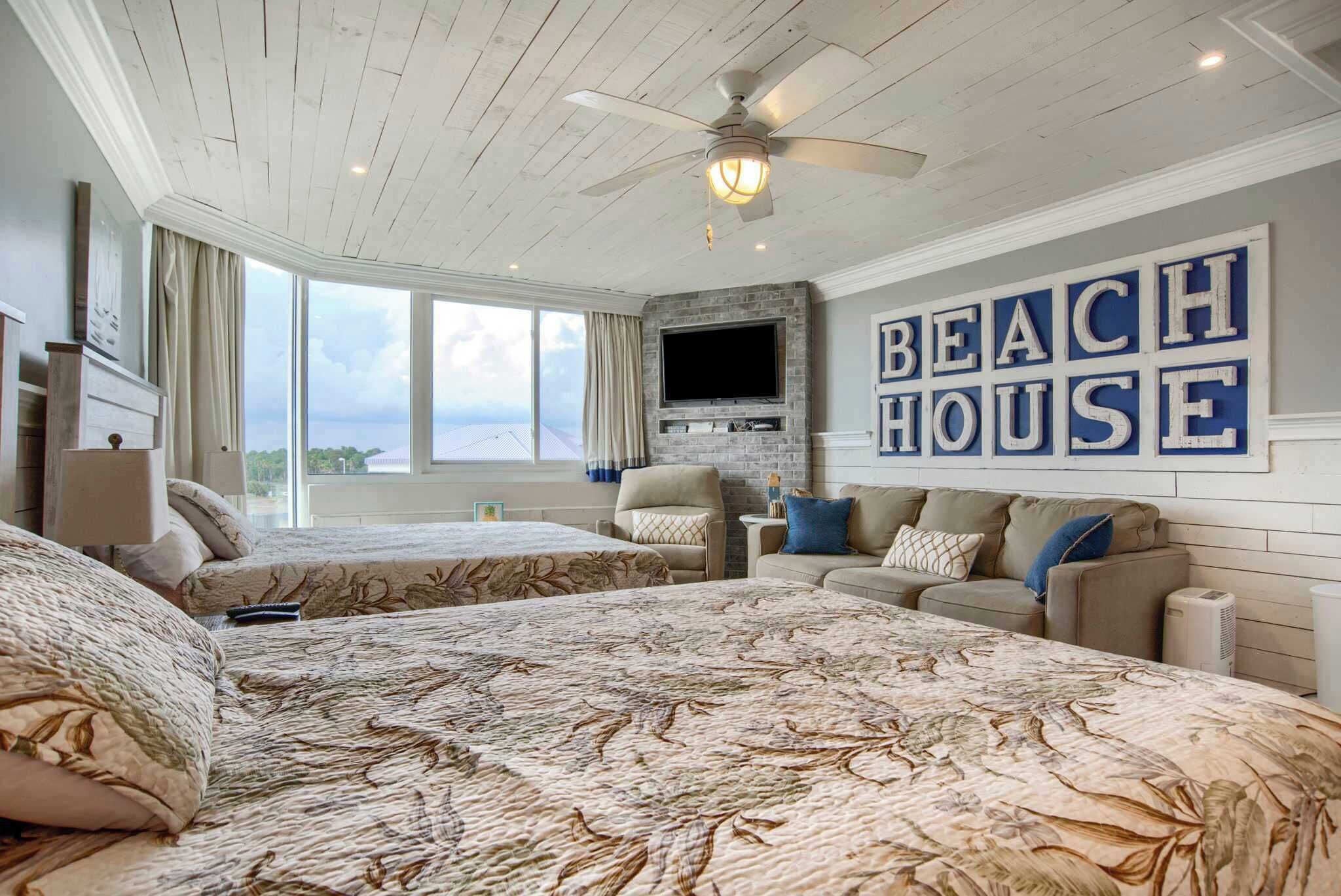 A bedroom with two beds, a ceiling fan, a TV mounted on a brick wall, and a large window offering a view.