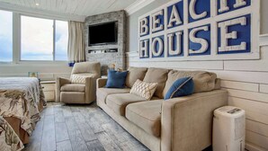 A living room with a beige sofa, a TV mounted on a stone wall, and a large 'BEACH HOUSE' sign on the wall.