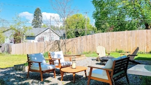 Ein Terrassenbereich im Freien mit Holzmöbeln, darunter Stühle und ein Tisch, umgeben von einem Holzzaun und Grünpflanzen.