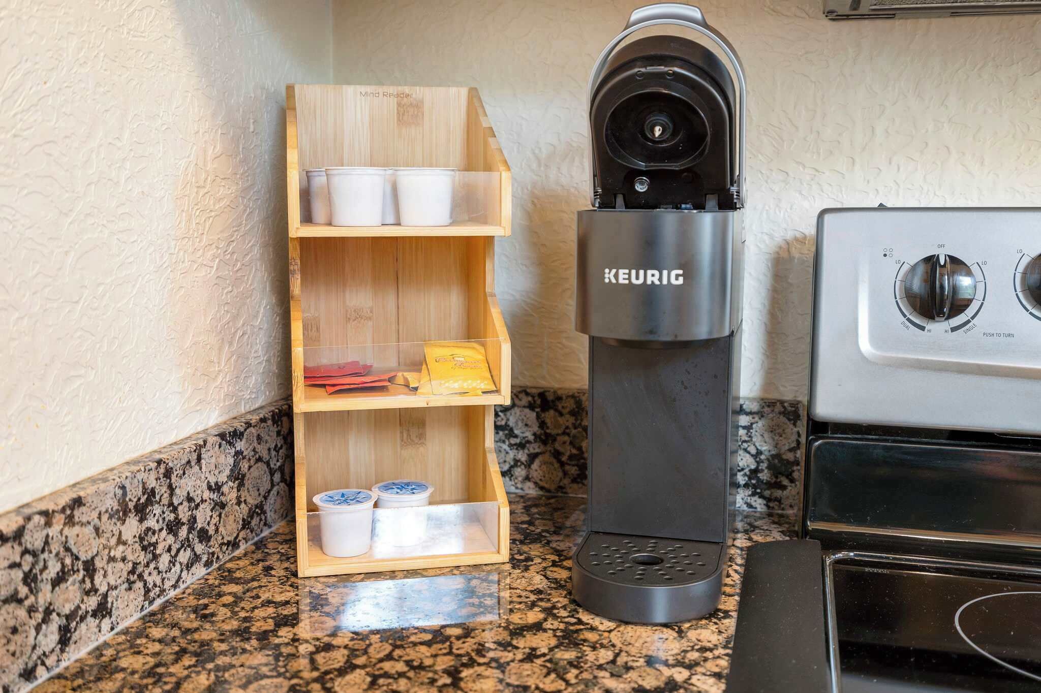 A kitchen with a Keurig coffee maker, a wooden shelf with cups and condiments, and a stove.