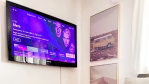 A living room with a flat-screen TV mounted on the wall, a white sofa, a small white table, and a vase with purple flowers. - Just Restored close to Franklin College and Lugano (Montagnola)