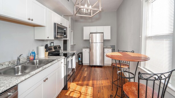 A modern kitchen with white cabinets, stainless steel appliances, a wooden floor, and a dining area with a table and chairs. - Suites on Main 3 (Covington)