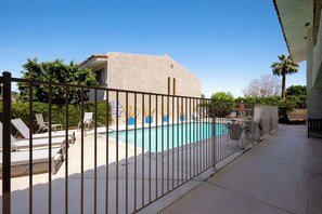 A swimming pool with lounge chairs and a fenced area, surrounded by trees and a building with a sloped roof. - Sunkist at 411 - Community Pool (Palm Springs)