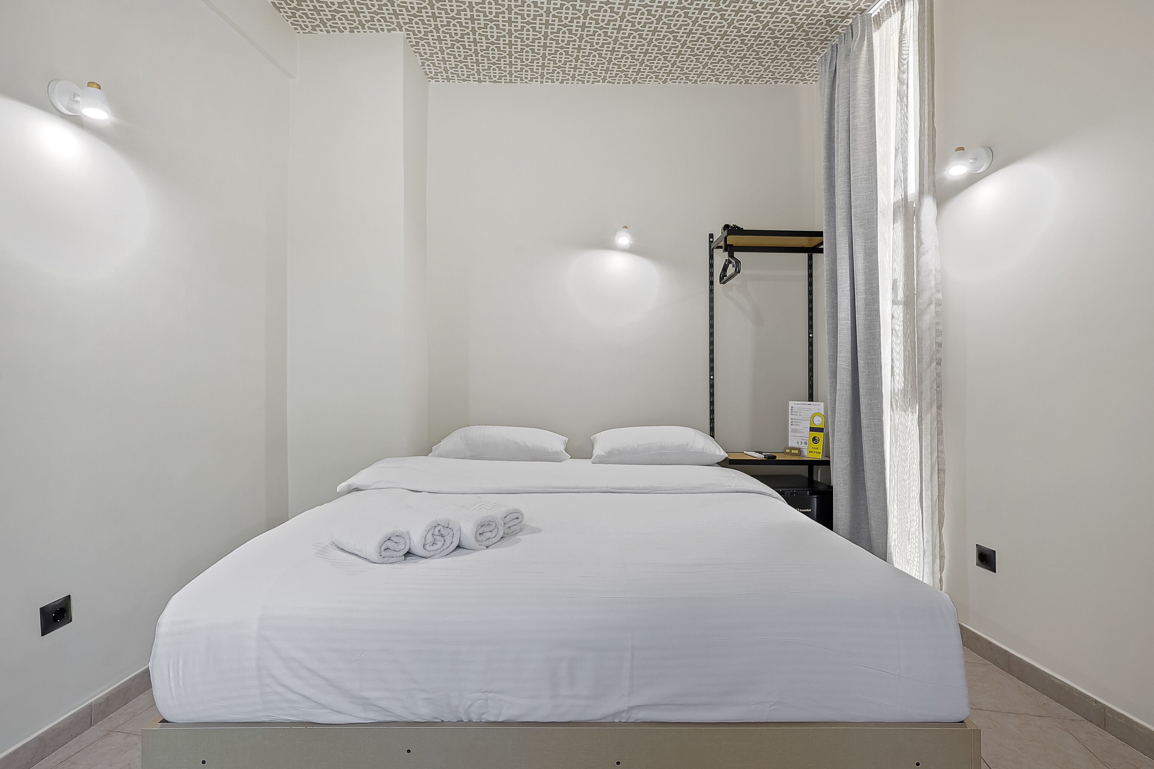 A neatly made bed with white linens and rolled towels, a bedside table with a lamp, and a wall-mounted shelf with a small item.