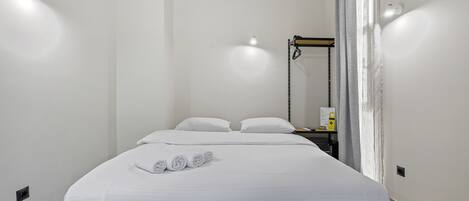 A neatly made bed with white linens and rolled towels, a bedside table with a lamp, and a wall-mounted shelf with a small item.