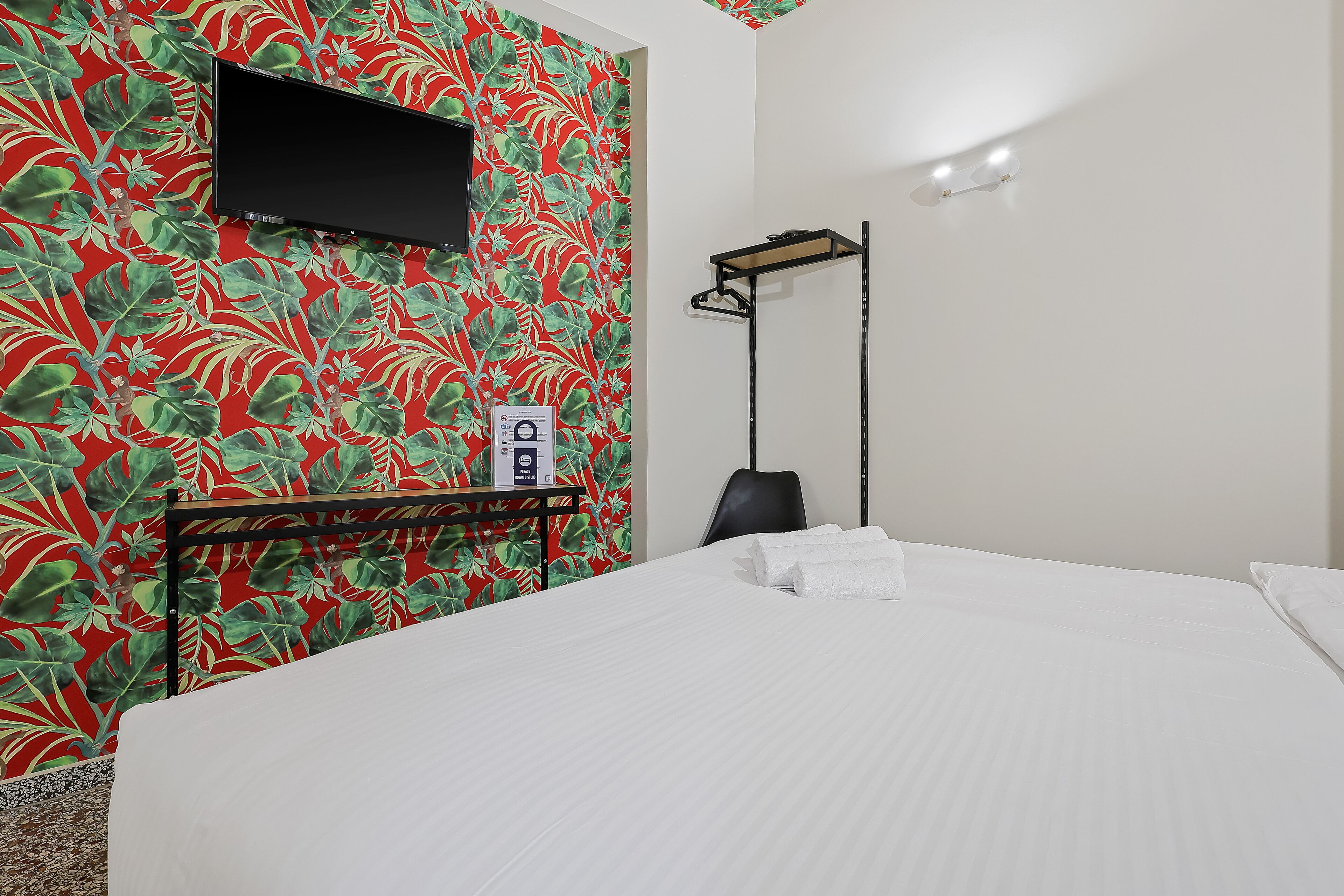 Una camera d'albergo con un letto, una TV a muro, una sedia nera e un tavolino con un asciugamano.