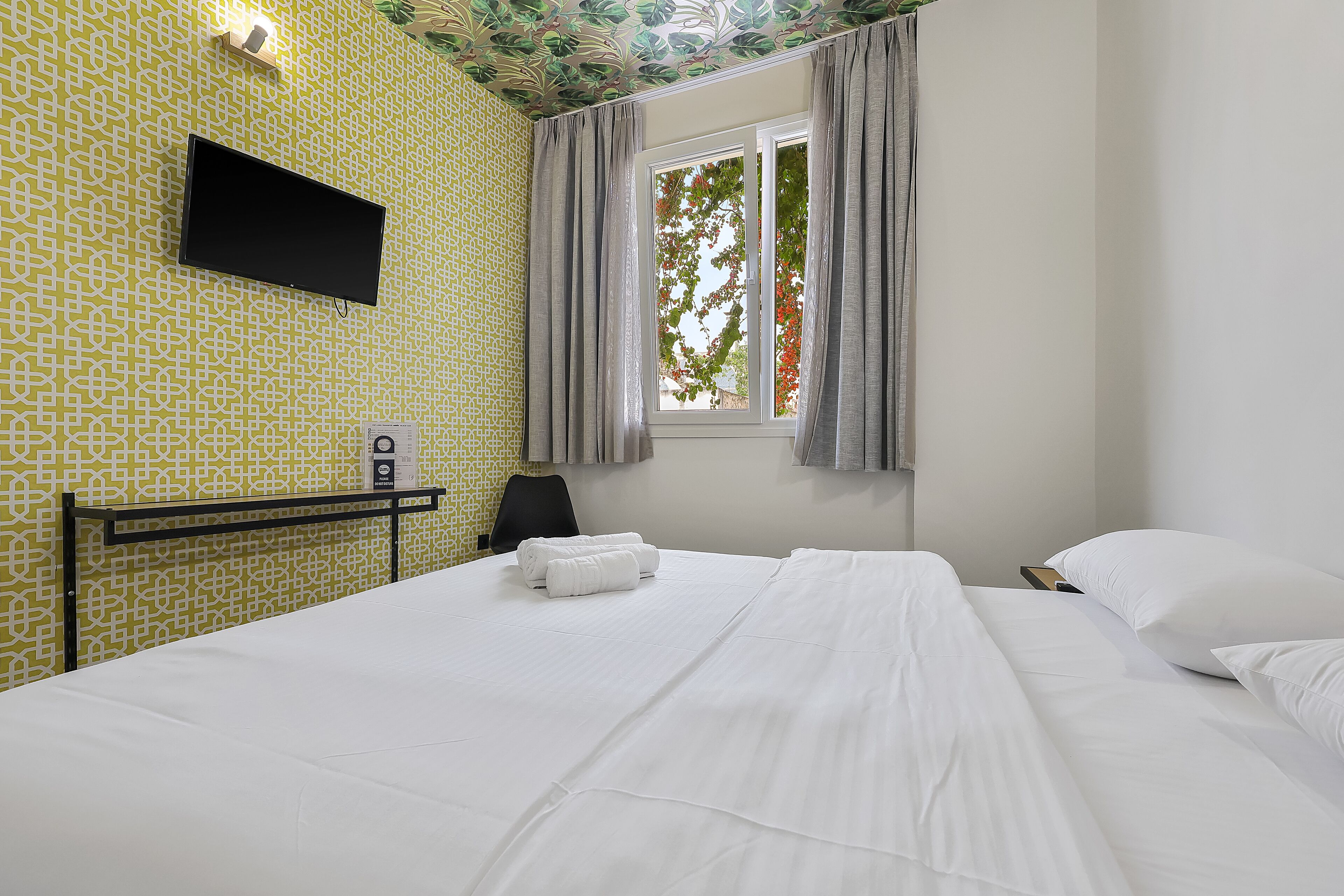 Ein Hotelzimmer mit gemustertem Wandpapier, einem an der Wand montierten Flachbildfernseher, einem Bett mit weißen Leinentüchern, einem Fenster mit Vorhängen und einem Schreibtisch mit Stuhl.