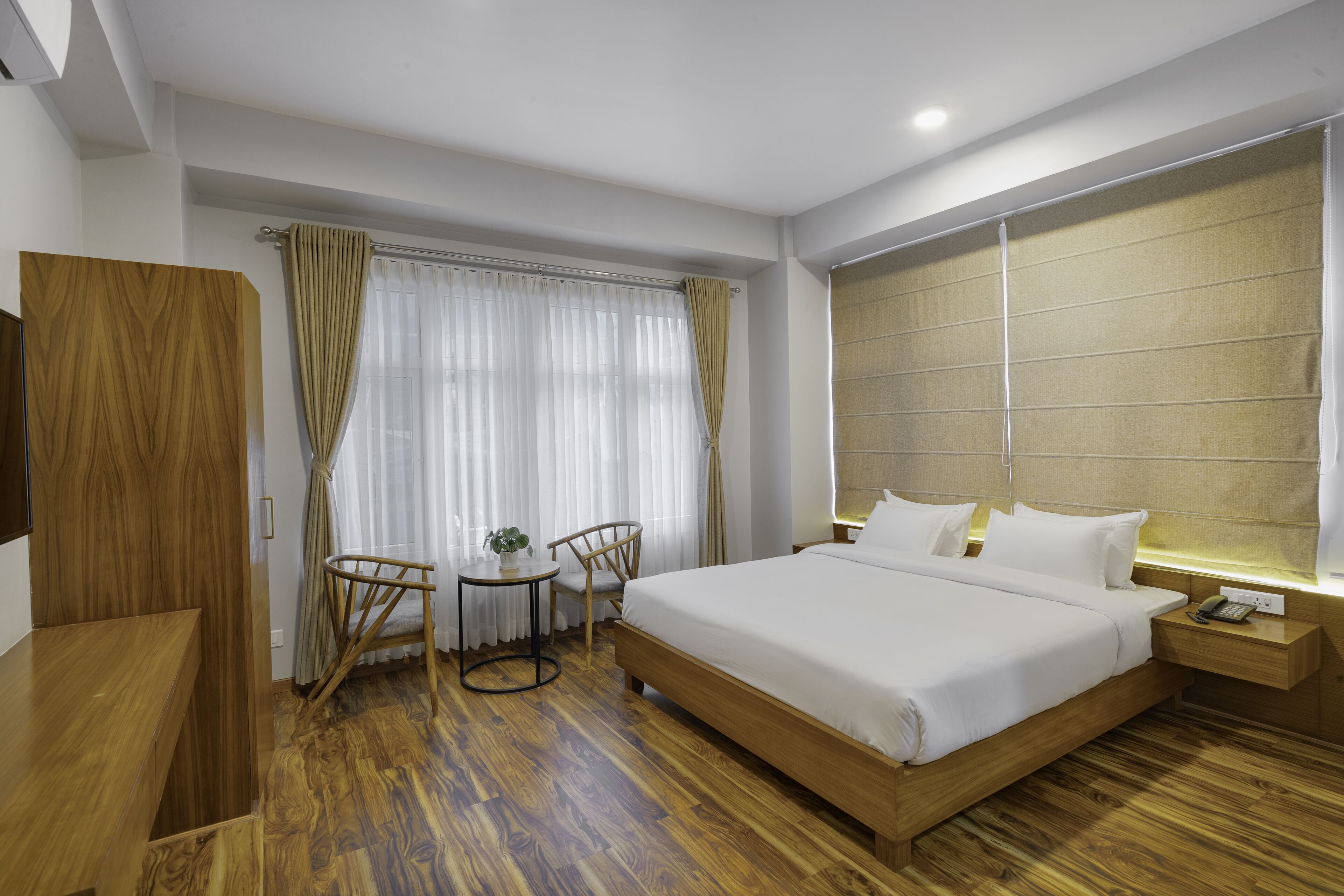 Ein Hotelzimmer mit einem Holzbett, einem Schreibtisch, einem Stuhl, einem kleinen Tisch mit Pflanze und einem Fenster mit Vorhängen.