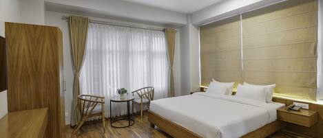 Ein Hotelzimmer mit einem Holzbett, einem Schreibtisch, einem Stuhl, einem kleinen Tisch mit Pflanze und einem Fenster mit Vorhängen.