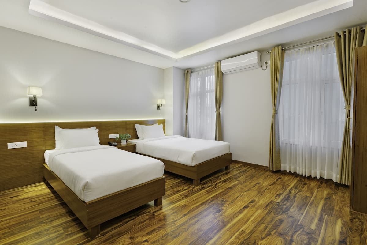 A hotel room with two beds, wooden flooring, a headboard, bedside lamps, an air conditioning unit, and curtains.