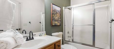 Un baño con lavamanos blanco, tocador de madera, inodoro blanco, cabina de ducha con mampara de cristal y un cuadro en la pared.