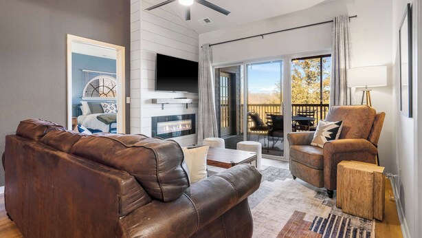 A modern living room with a leather sofa, a fireplace, a ceiling fan, and a large window with a view of trees. - T Time Getaway - Overlooking Golf Views! (Branson West)