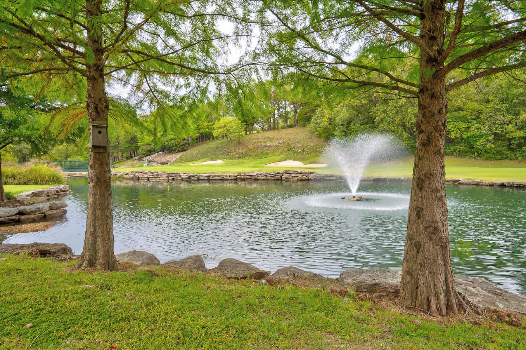 A tranquil pond with a fountain, surrounded by lush greenery and trees.