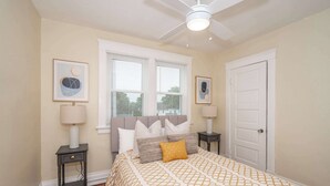 Une chambre à coucher comprenant un lit, deux tables de chevet, un ventilateur de plafond et deux fenêtres équipées de stores.