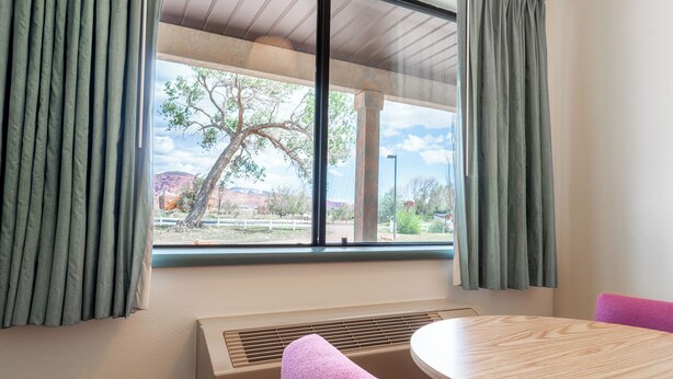 Una habitación con un ventanal grande, una mesa redonda de madera, un banco con cojín rosa y un respiradero color beige.