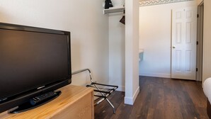 Una habitación con una televisión de pantalla plana sobre un soporte de madera, un atril metálico, una puerta blanca y un pasillo con suelo de madera.