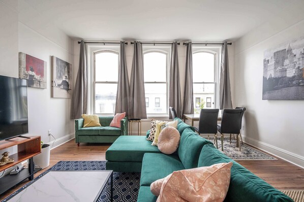 A living room with a green sofa, a TV, and a dining area with chairs. - The Heart of Madison 3 (Covington)