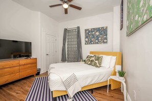 Une chambre à coucher comprenant un lit, une commode en bois, un téléviseur, un ventilateur de plafond et des œuvres d’art accrochées aux murs.