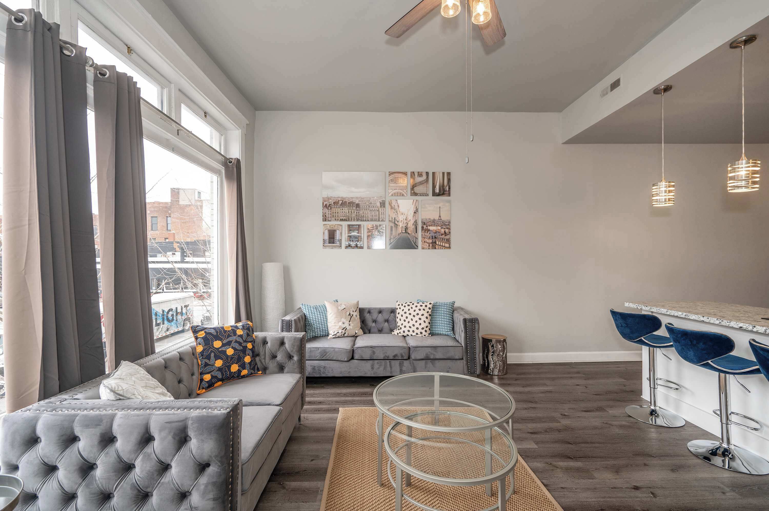 A modern living room with a grey sofa, a glass coffee table, and a bar area with blue bar stools.