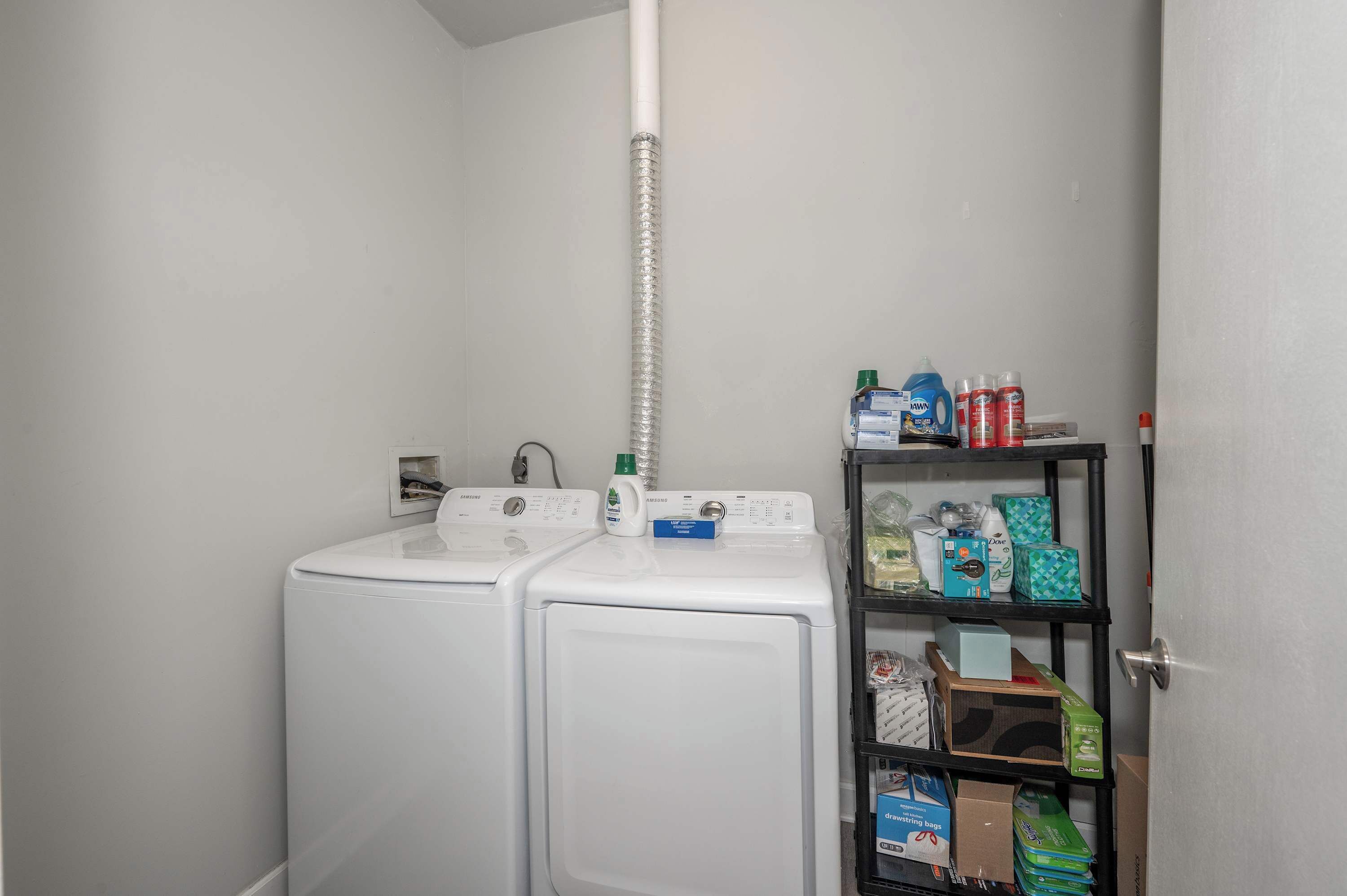 A laundry room with two white front-loading washing machines, a metal vent pipe, and a shelf stocked with cleaning supplies.