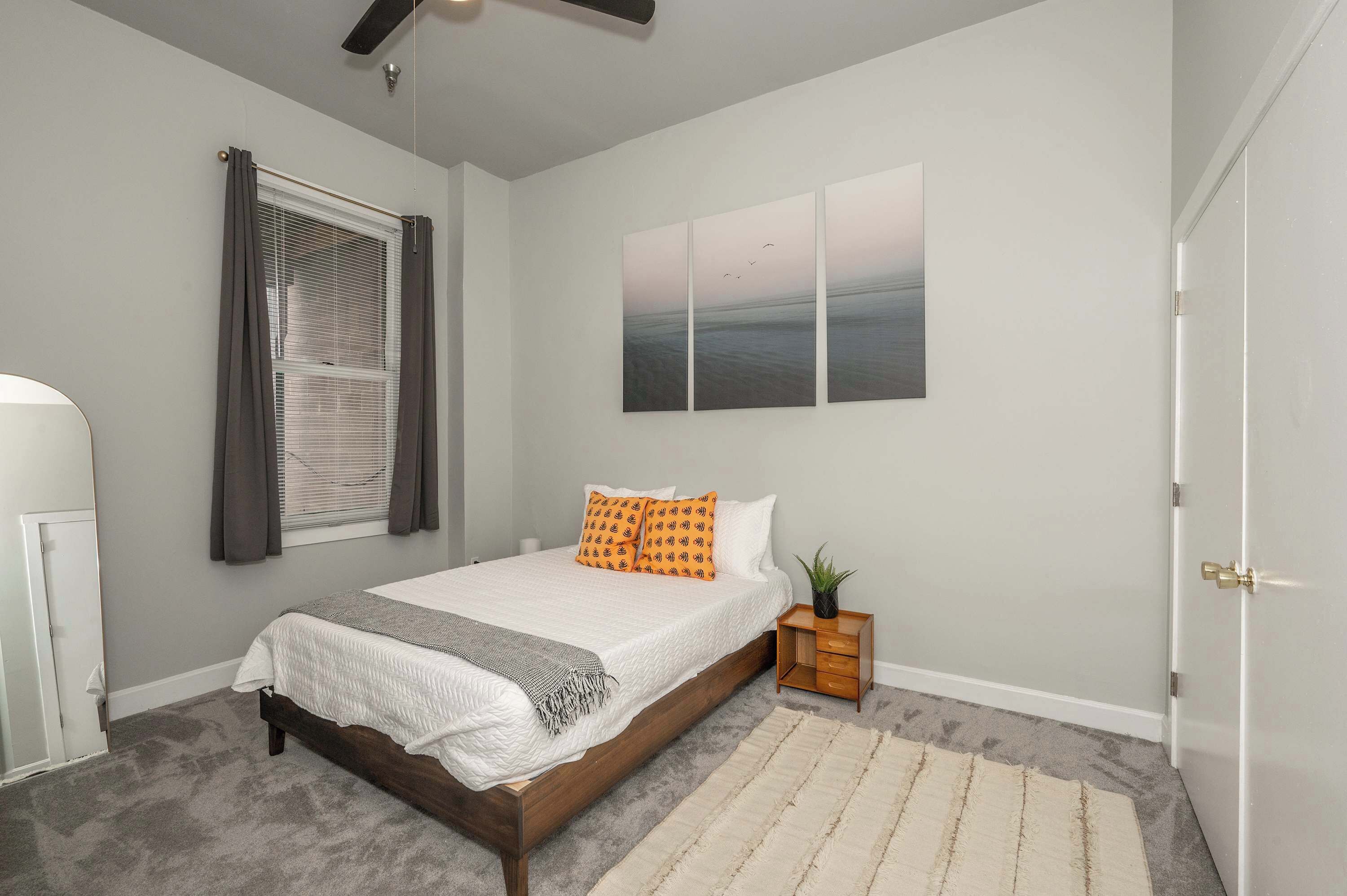 A bedroom with a bed, a nightstand, a window with curtains, a wall-mounted artwork, and a door.