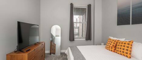 A bedroom with a TV on a wooden dresser, a bed with white bedding and orange pillows, a window with curtains, and a painting on the wall.