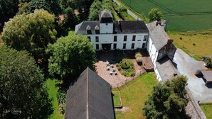 An aerial view of a large, white building with a symmetrical facade, surrounded by lush greenery and a well-maintained courtyard with outdoor seating.
