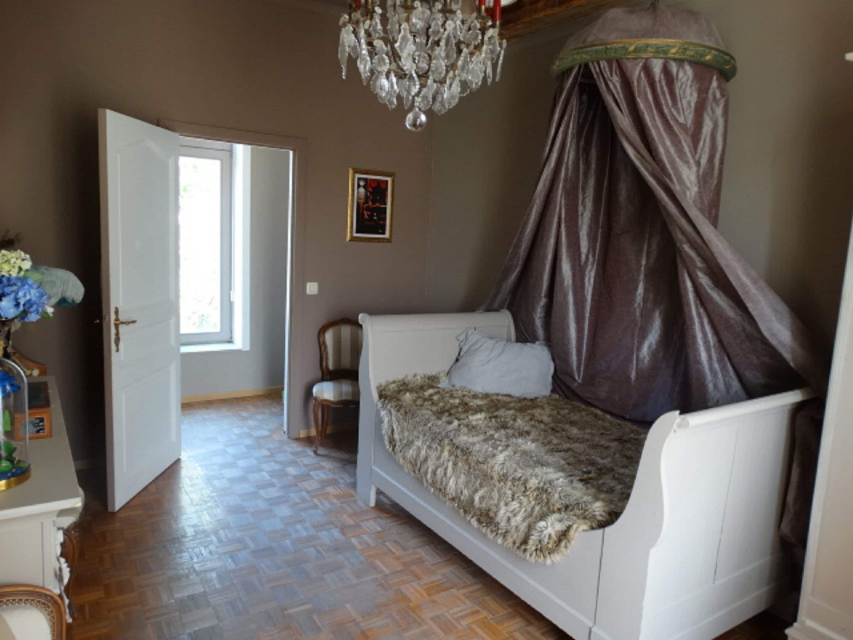 A bedroom with a canopy bed, a chandelier, a chair, and a door leading to another room.