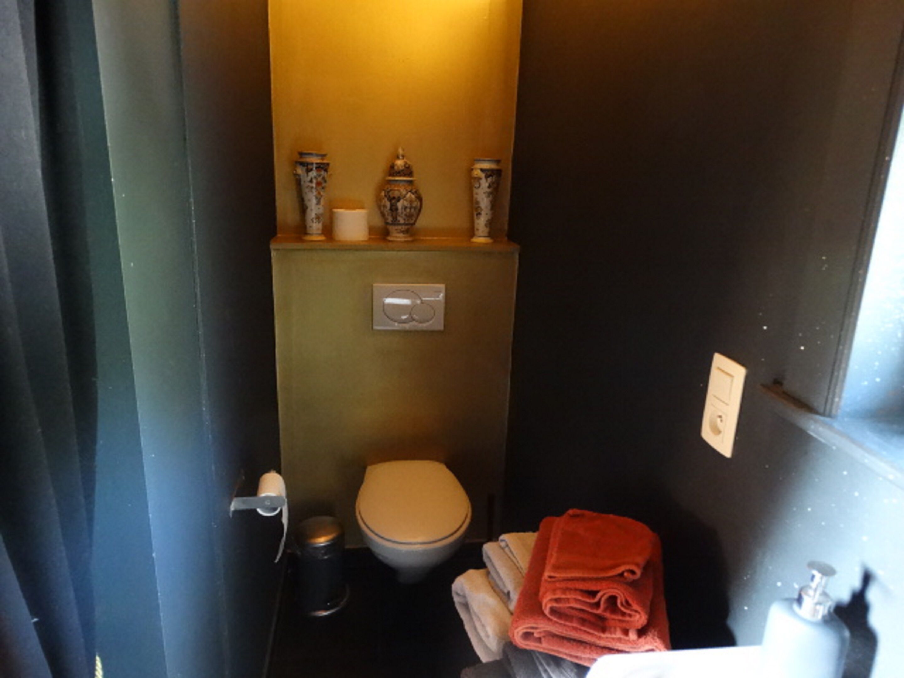 A small, narrow bathroom with a toilet, a trash can, and a stack of towels.