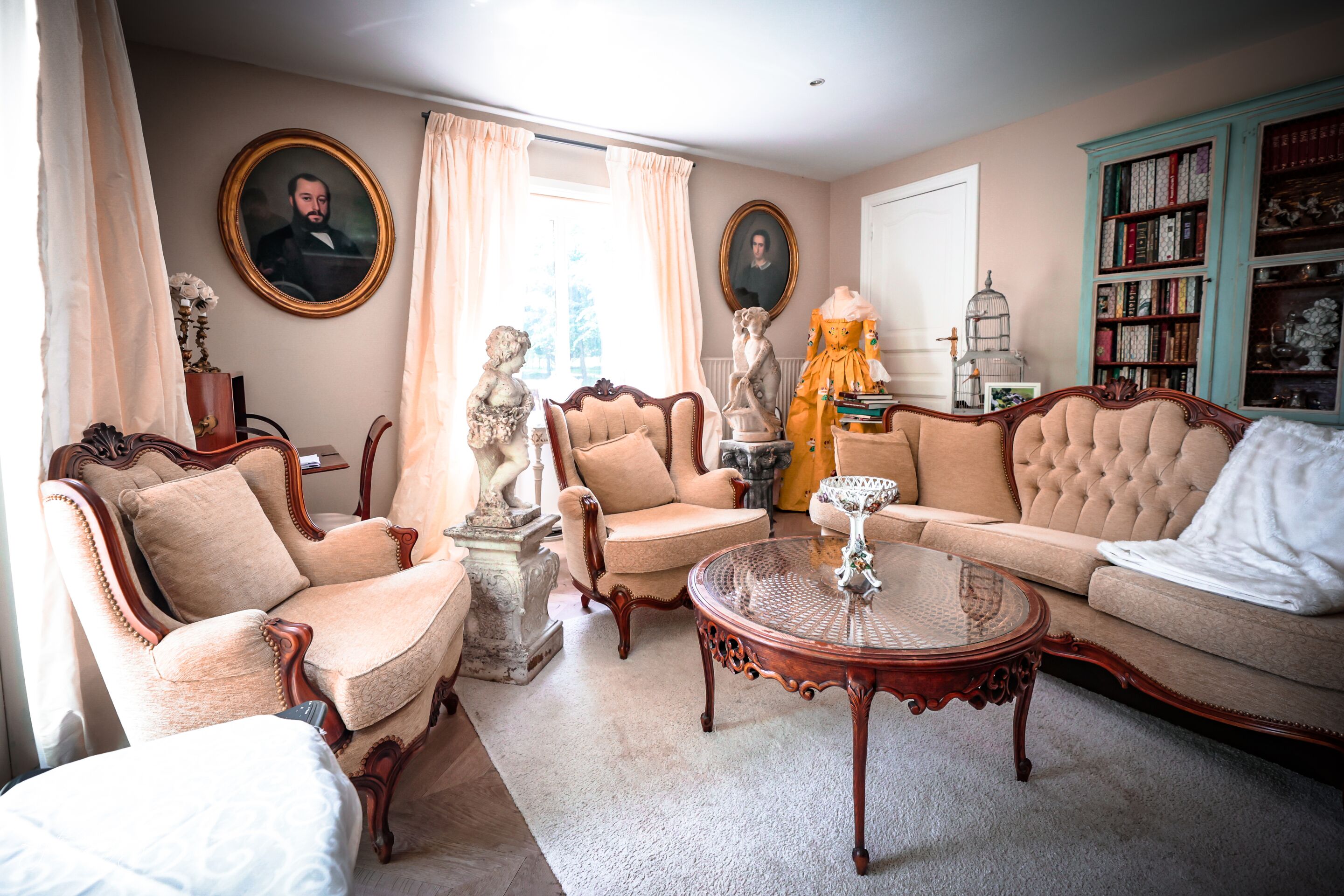 Un salón clásico con muebles antiguos, incluyendo un sofá, sillas y una mesa de centro. Hay dos cuadros en las paredes y una estantería llena de libros.