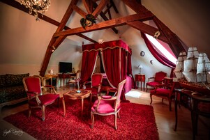 Una habitación con techo alto, vigas de madera, alfombra roja y una cama con dosel. Hay varias sillas y una mesa con lámpara.