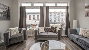 A living room with two grey sofas, a glass coffee table, and a view of buildings through large windows. - The Heart of Madison Suites (Covington)