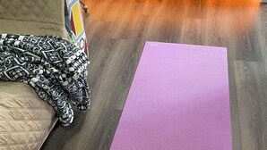 Quarto com piso de madeira, cama com cobertura acolchoada, mesa de vidro com abajur e tapete de exercícios roxo.