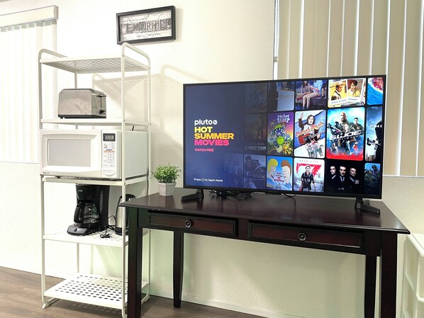 A flat-screen TV displaying a selection of summer movies on Pluto TV, a white shelf unit with a microwave, a coffee maker, and a small potted plant.