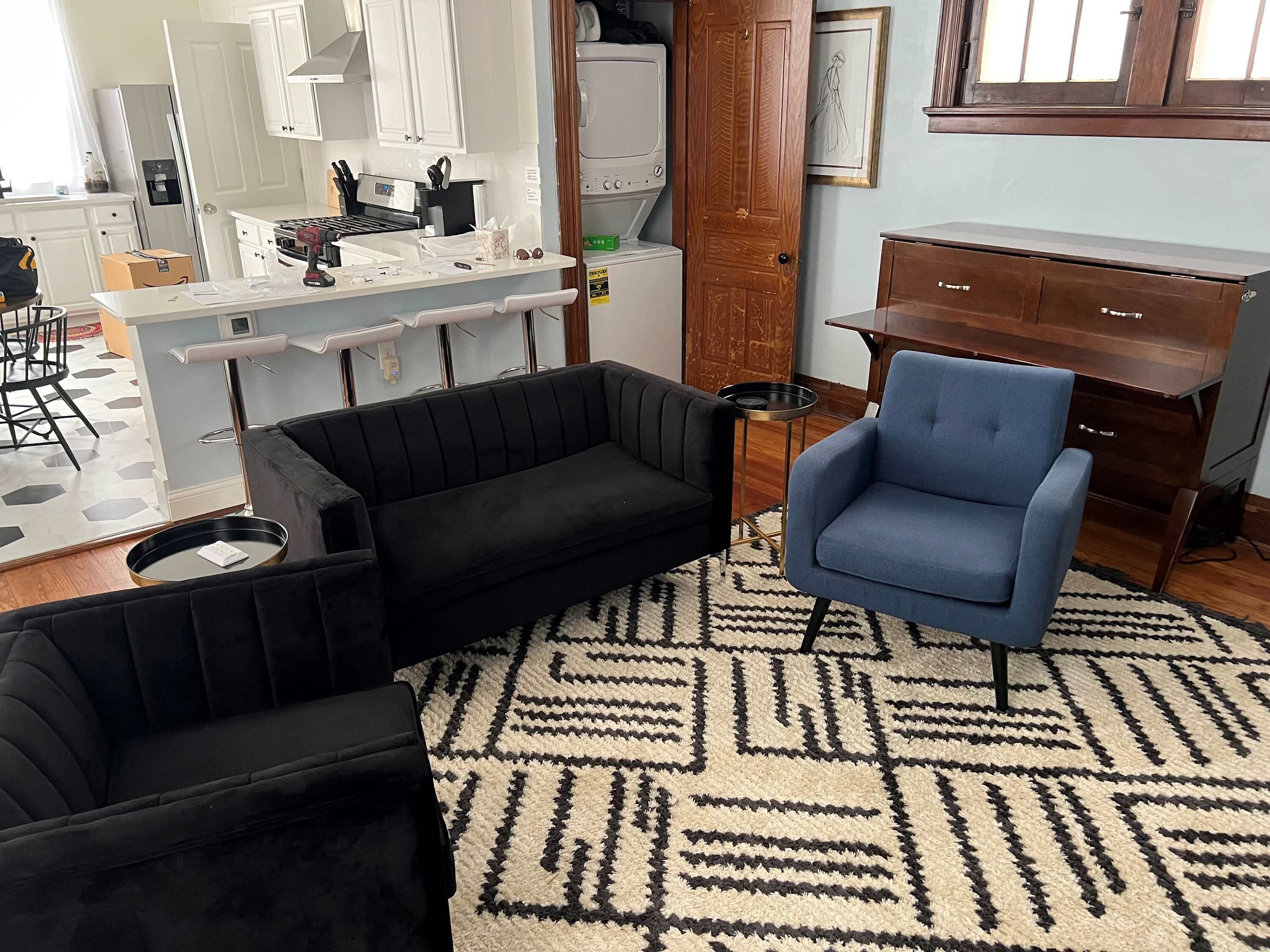 Una sala de estar con un sofá negro, un sillón azul y un tapete estampado.