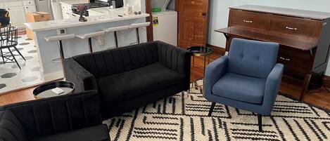 Una sala de estar con un sofá negro, un sillón azul y un tapete estampado.