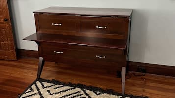A wooden dresser with drawers against a wall, a window above, and a patterned rug on the floor.