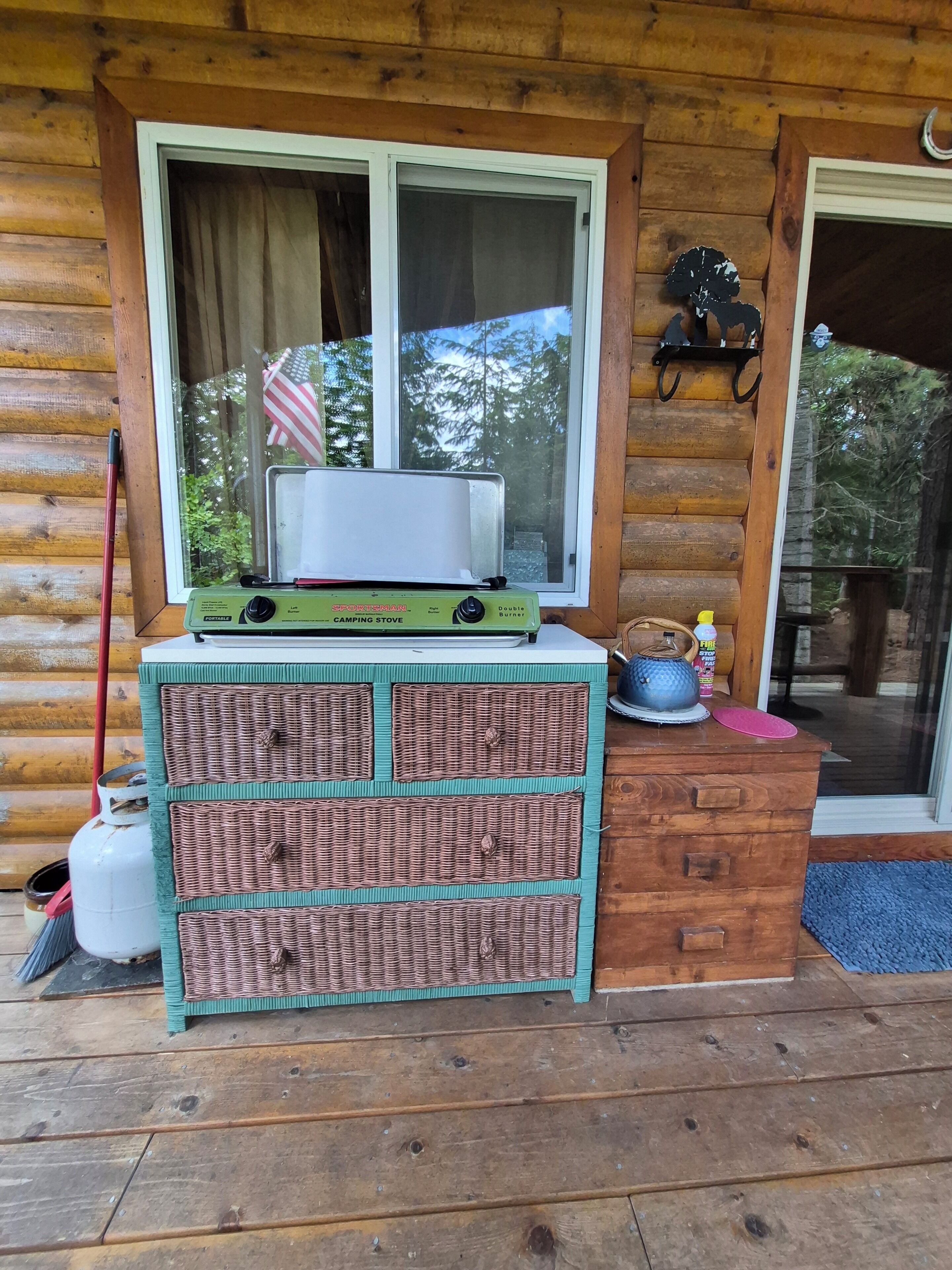 A wooden cabin with a window, a portable camping stove, a wicker storage unit, and a gas canister.