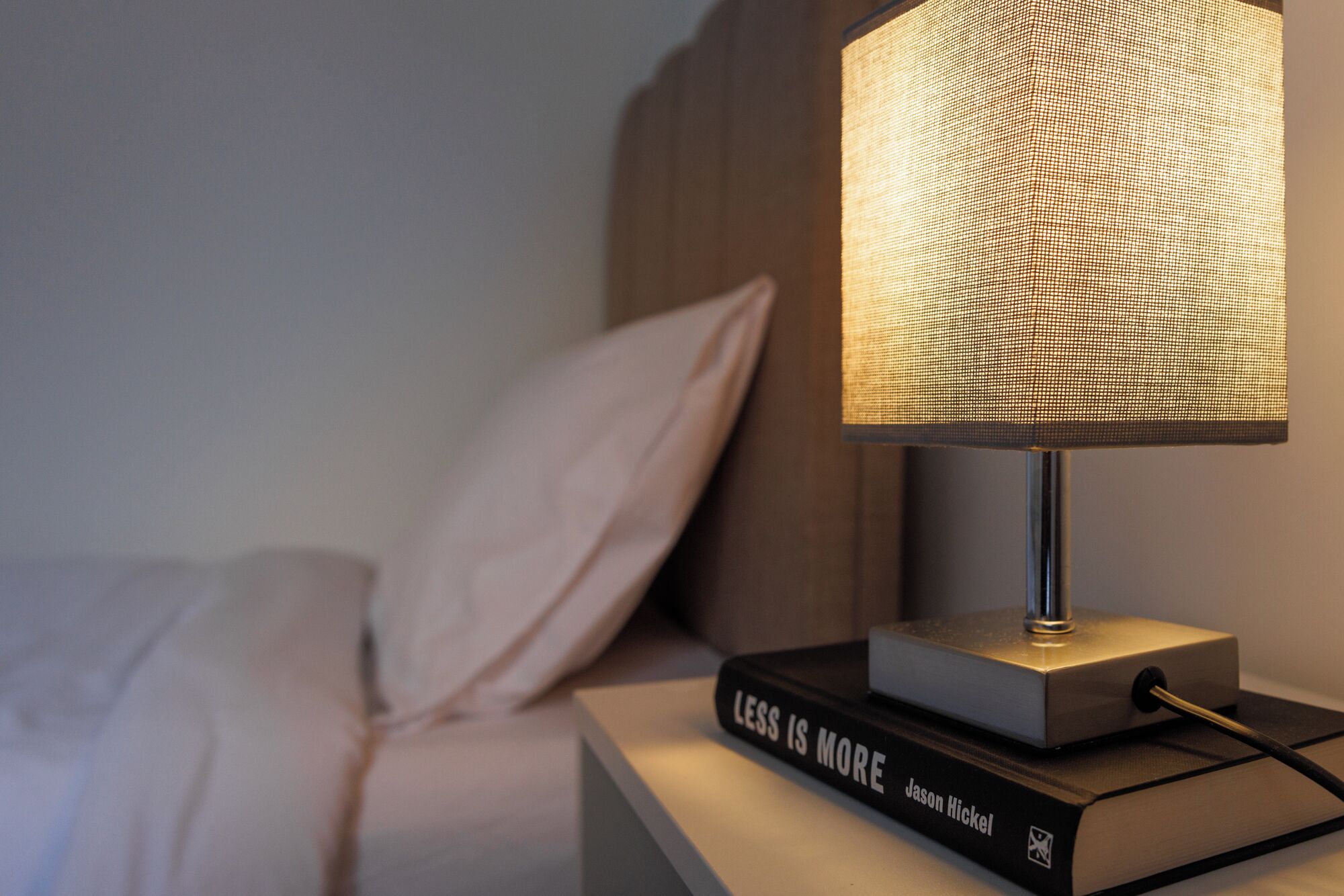 Ein Nachttisch mit Lampe und dem Buch 'Less is More' von Jason Hickel.