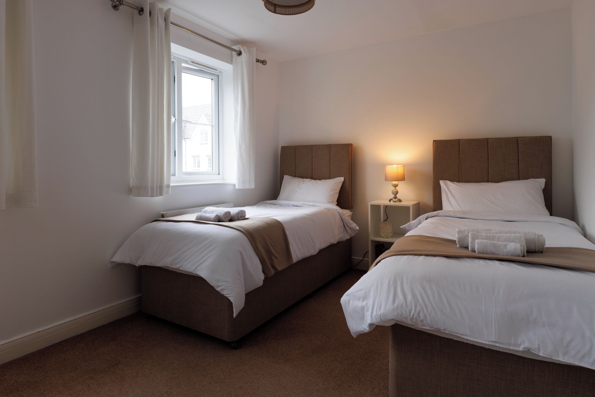 Een hotelkamer met twee bedden, een raam met gordijnen, een bedlampje en een klein plankje.