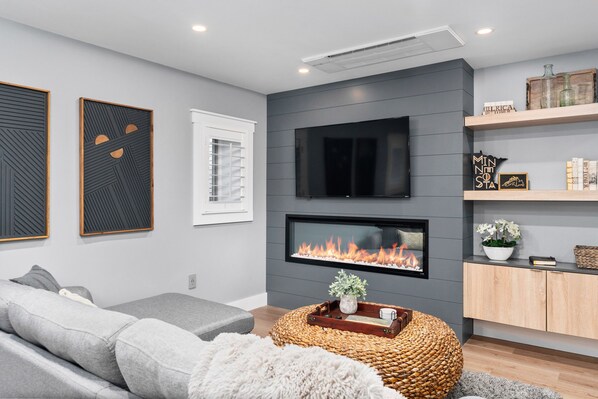 Un salon moderne avec un canapé gris, un téléviseur à écran plat fixé au-dessus d’une cheminée et un meuble en bois agrémenté d’objets décoratifs.
